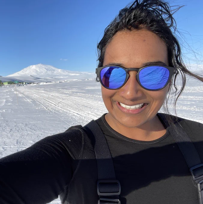 Maria, with reflective sunglasses, stands on the sea ice in front of Mount Erebus, with part of Scott Base in the background.