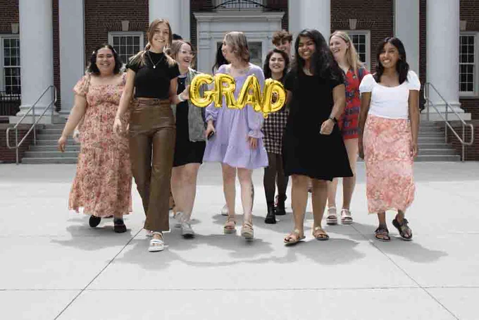 Twelve students walk in front of UNO's Arts and Science Hall. They are holding a mylar balloon that spells out grad.