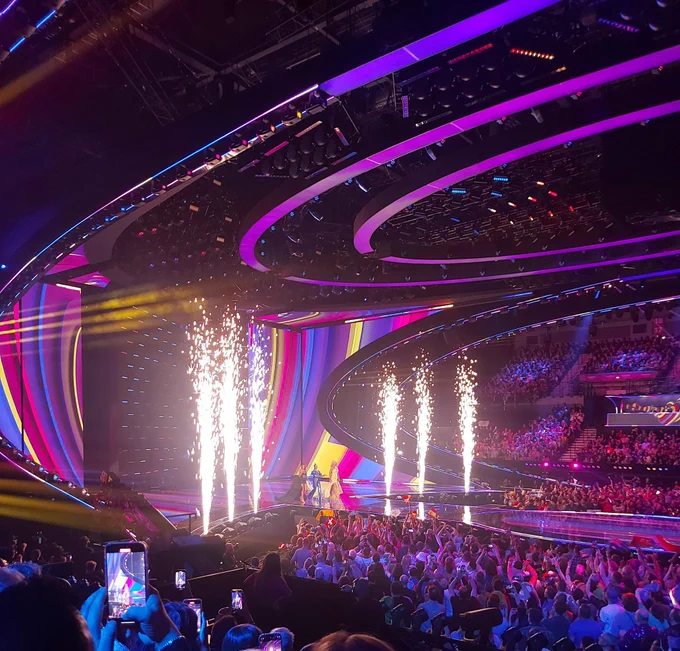 The three hosts (left-to-right, Alesha Dixon, Julia and Hannah Waddington) appearing onstage at the Eurovision Song Contest Semi Final 2. They are surrounding by lighting in pink and blue with six pyrotechnics.