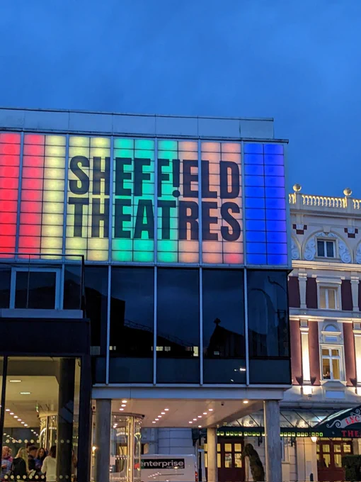 SHEFF!ELD THEATRES brand signage lit up in multiple colours above the crucible with folks in foyer and the lyceum building also in the background