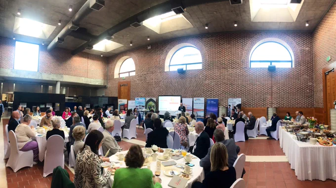 Guests pictured in the Dining Hall, Tourism and Hospitality Building MTU. Guests are enjoying breakfast, are sitting at round tables. A female speaker is standing at the top of the room with a screen and pull up signs behind her.