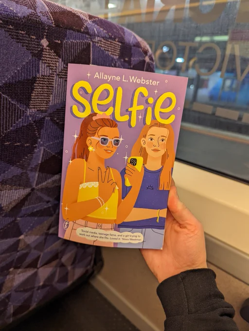 Hand holding up book on train: Selfie by Allayne L. Webster