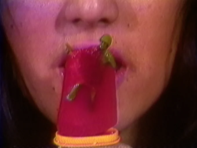 A video still from one of Chile's first female videographers, Gloria Gloria Camiruaga. It depicts a close-up of the lower half of a woman's face. She holds a red popsicle with a green plastic toy soldier embedded in it to her mouth. 