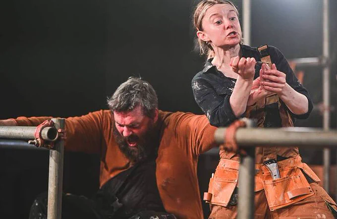 A performer in a brown shirt holding into two metal posts with his mouth open, looking as if he is in pain. Another performer stands next to him and is holding something against his finger