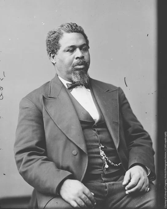 A black and white portrait of Robert Smalls. He is seated wearing a formal three three piece suit with a chain connected a button and a black bowtie. 