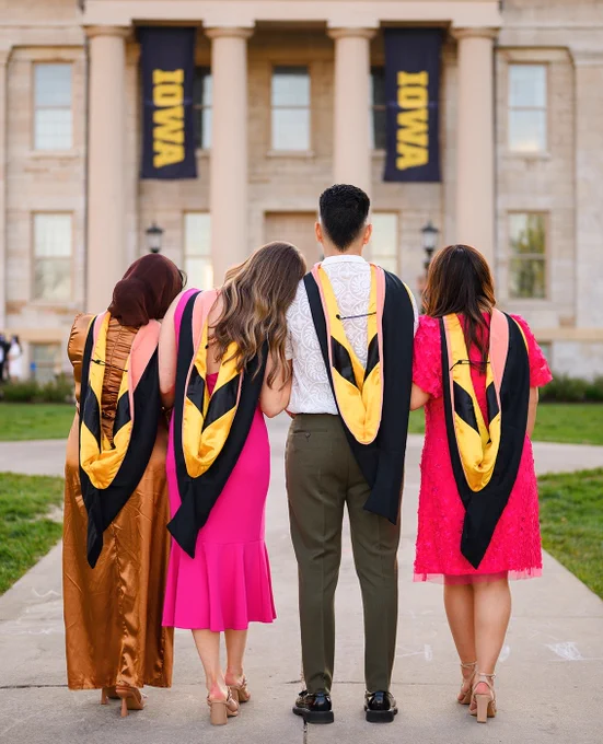 4 graduating students, backs to the camera, facing Old Capitol with black and gold IOWA banners hanging on the portico.