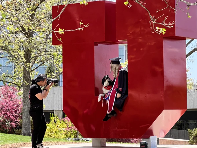 Photo of photographer taking a photo of a graduate in her regalia sitting on the Block U on campus with her dog next to her.