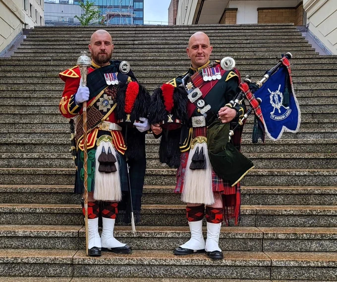Two male identical twin British soldiers are standing, facing the camera, on some stone steps.

The soldier on the left is holding a drum majors mace upright in his right hand and a black bonnet with a red plume in his left hand.  He is wearing a red jacket with gold shoulder wings and gold coloured rank on his arm, and a Drum Majors cross belt. He is also wearing a Black Watch tartan kilt with a whited haired, black tassled sporran.  He has red and black stockings with white gaitors over his shoes.

The soldier on the right is holding a set of bacg pipes in his left hand and a black bonnet with a red plume in his right. He is wearing a green jacket with gold shoulder wings and piping, and a black cross belt across his body. He is also wearing a Royal Stuart tartan kilt with a whited haired, black tassled sporran and a Royal Stuart Plaid across his body, hanging from his left shoulder. He has red and black stockings with white gaitors over his shoes.