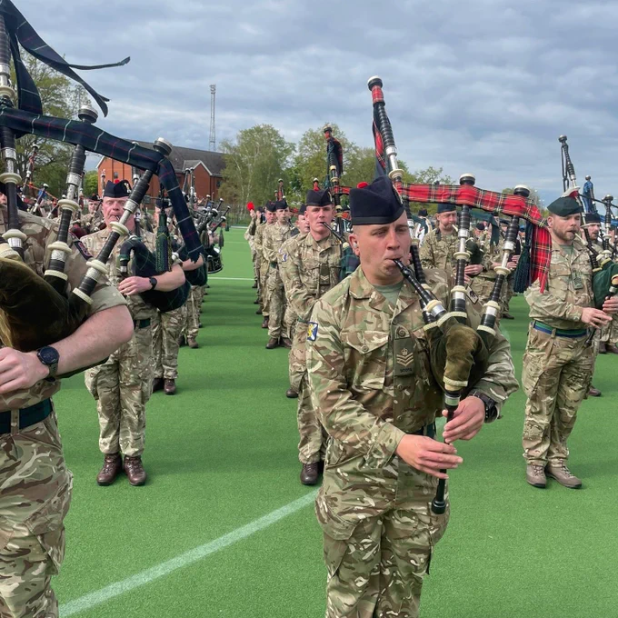 A group of British soldiers are standing in lines on green Astroturf, playing bagpipes.

They are wearing camouflage uniforms, with dark blue Glengarry’s on their heads.