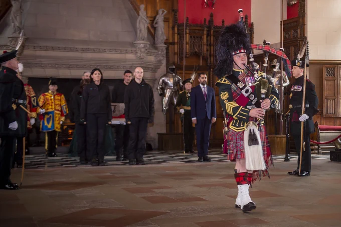 A British soldier, playing the bagpipes is marching across room with a stone covered floor. Behind him are four people, dressed in black clothing, carrying the Stone of Destiny. Either side of them are members of the Scottish Government, as well as two members of the Royal Company of Archers dresses in green uniforms with feathers in their hats.