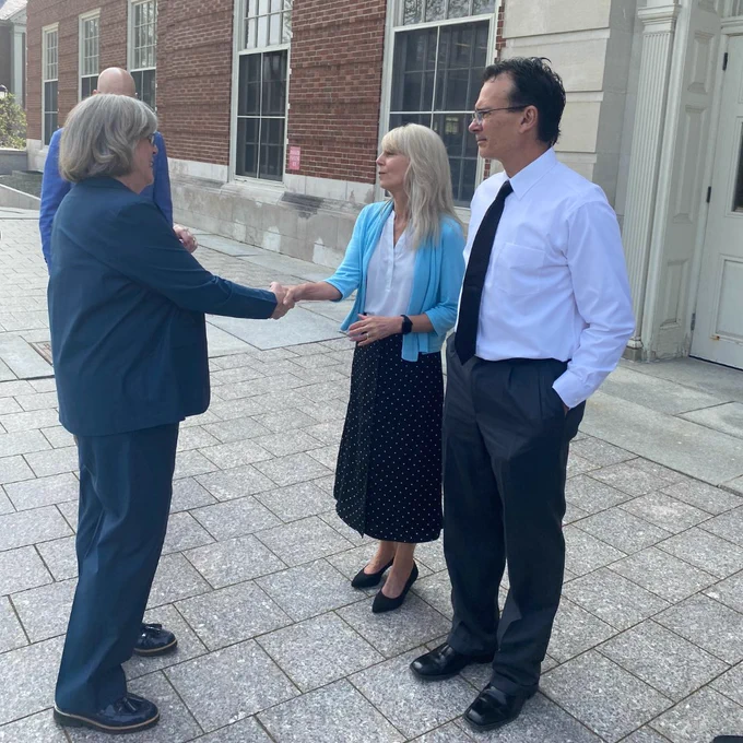 A photo of three people, two of which are shaking hands, standing in front of Fogler Library. 