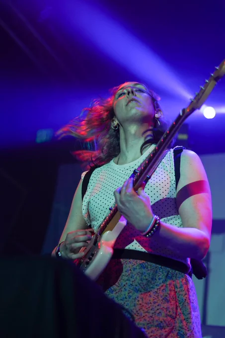 Portrait orientation photograph of nmlstyl. She is mid-guitar strum and illuminated by pink and purple stage lights, and her hair flowing to the right. 