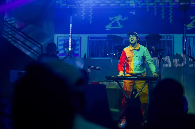 A photograph of Button Masher from the crowd. There are two out of focus figures in front of the photographer which provides a nice focal point to Jake, who is adorned with a classic snapback and a cool bicolor button-up jacket. His mouth is open because he's nailing a juicy piano chord, and behind him are projection-mapped visuals with his visualist Dæmon Core on stage. 