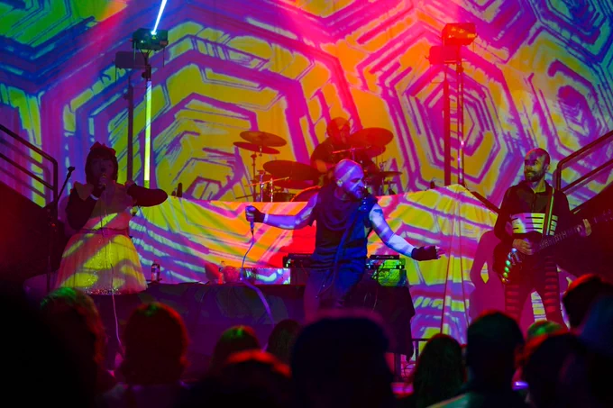 A psychedelic photograph of the full crashfaster band from the crowd. The visuals (courtesy of Dæmon Core) feature tiled lime green and magenta geometric shapes. Their drummer Devin is on riser, and vocalists Keiko, Morgan, and guitarist Ryan on the stage. 