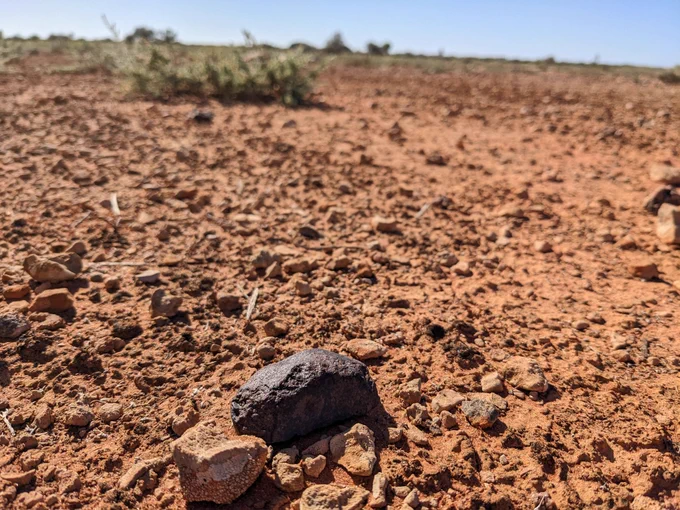 A dark stone sat in the foreground of a landscape photo, with a red sandy ground surface covered in small red coloured rocks
