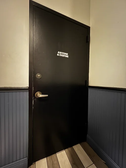 A black door leading to a single toilet. The sign on the door says ‘anyone & everyone’ in white writing.