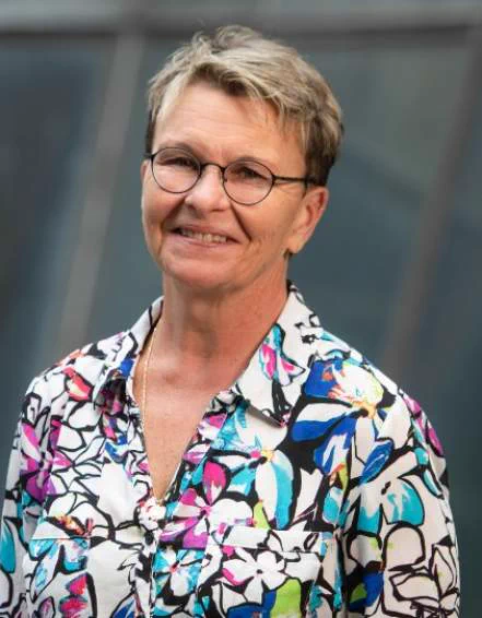 Susan Green is a White woman in her fifties, wearing short blonde-brown hair, glasses, and a multi-colored shirt with large abstract floral print.