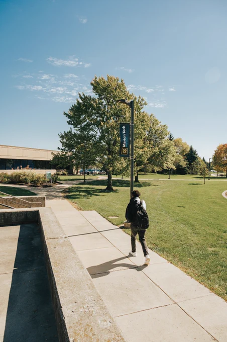 A student with a backpack walks across campus on a sunny day, with green grass, green trees, and a Marion Tech banner on a light pole. 