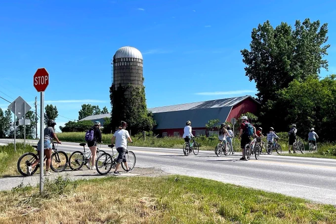 Children wearing helmets walk their bikes across a Vermont road, there is a red barn and a silo in the background. 
