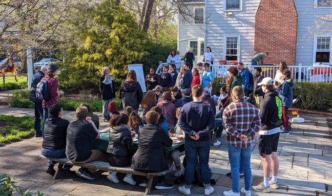 The consul general addresses group of students and faculty at URI. The group is sitting on picnic tables and standing.