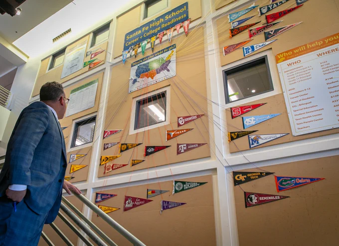 A photo of Secretary Cardona looking up at a wall of college pennants with strings leading to each of their locations on a map of the United States.