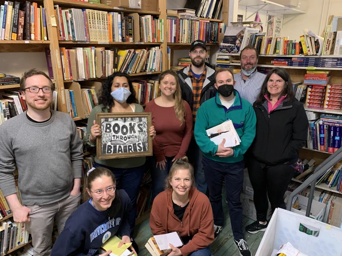 9 GPLLA members and friends stand in front of bookshelves at Books Through Bars. One person holds up a "Books Through Bars" sign.