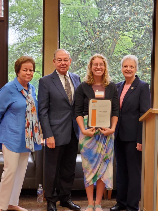 4 people stand in front of a window with trees outside; one holds her certificate