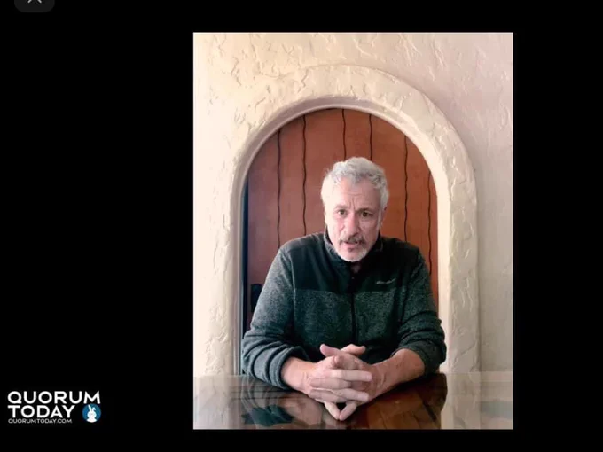 “Q” is shown in an image from the video on QuorumToday.com. His fingers are intertwined and Q faces the camera intently.  The background, which includes an arched doorway, and Q’s image reflects on the counter.