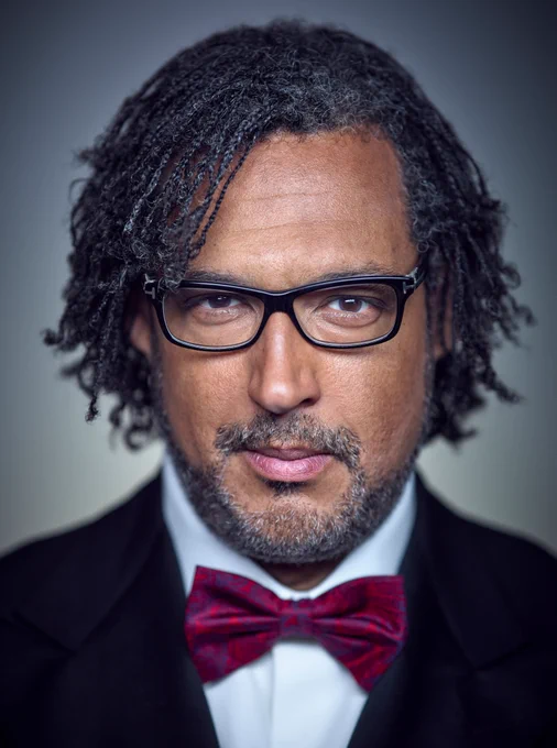 A close up portrait of Professor David Olusoga OBE. He is wearing black framed glasses and a black suit and a maroon bowtie.