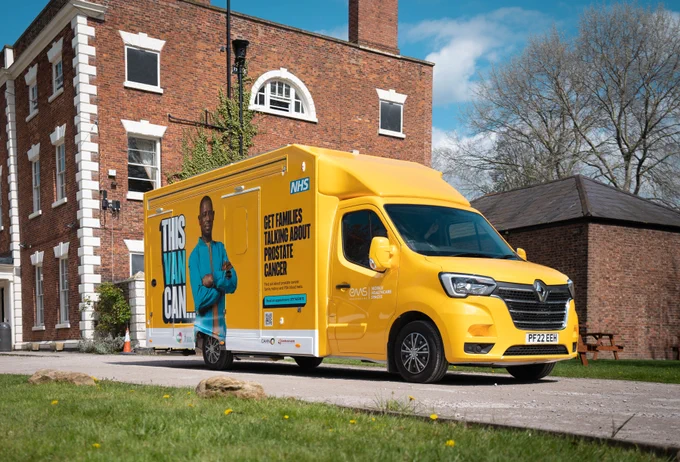 A yellow van with the words 'ThisVanCan get families talking about prostate cancer' on the side and an image of Gilly, a black man wearing a blue top drives along a road.