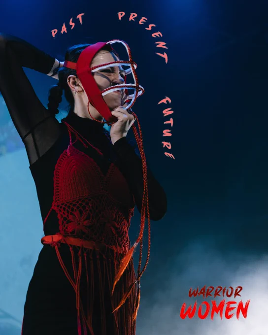  a close up of a female performer wearing a crochet costume & mask with her hand on the front and back of the mask with the text "past present future" overlaid and the text "warrior women" in the bottom right corner
