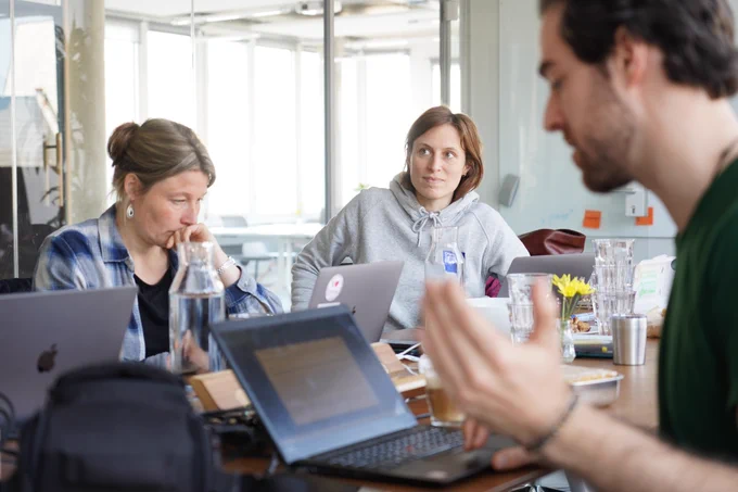 Zwei Teammitglieder hören einem dritten Teammitglied gespannt zu. Alle sitzen um einen großen Konferenztisch. 