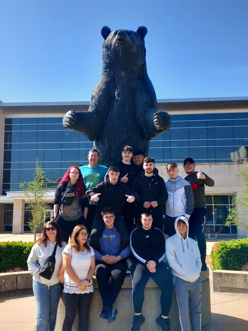 13 students posing for a photo in front of a bear statue on a sunny day in Missouri State University.