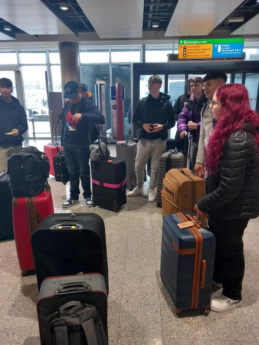 Students stood around waiting with their suitcases in the airport. There are 7 students visable.