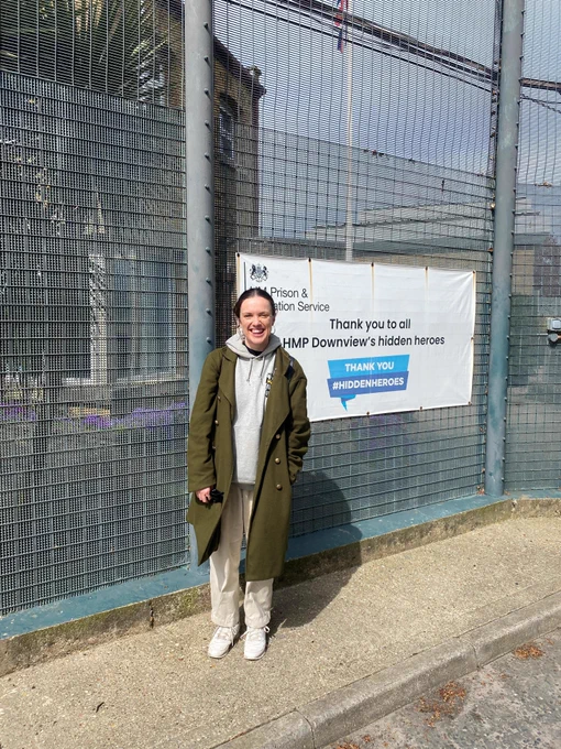 El, Director of Kestrel Theatre, standing outside HMP Downview and smiling to camera. El is wearing a long brown coat over a cream outfit.