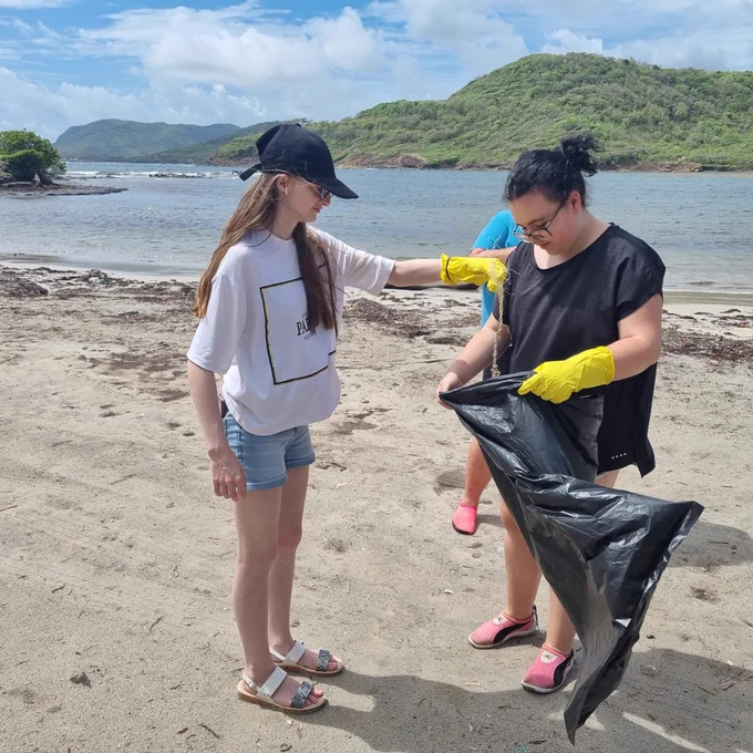 A photo of 2 students picking up litter on the beach and placing it into a black bin bag. The students are wearing yellow rubber gloves.