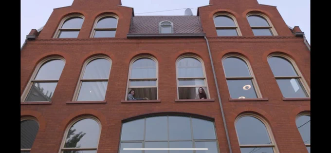 Front of a building with two open windows next to each other on the second floor. A man is in the one on the left and a woman is in the one on the right. The building is brick and a very nice house.