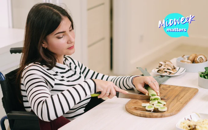 Lady with long dark hair sitting in a wheelchair at a table cutting up vegetables on a wooden chopping board. using a knife in her right hand and holding a leak in her left hand wich is resting on the chopping board.