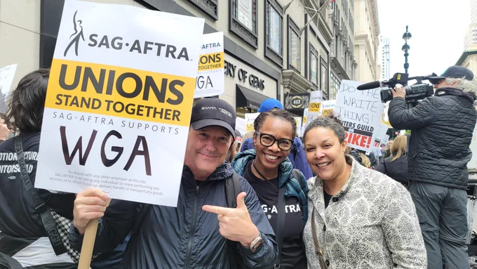 Clark Gregg, Avis Boone, Linda Powell. Clark holds a sign that reads 'SAG-AFTRA Unions Stand Together. SAG-AFTRA supports WGA.'