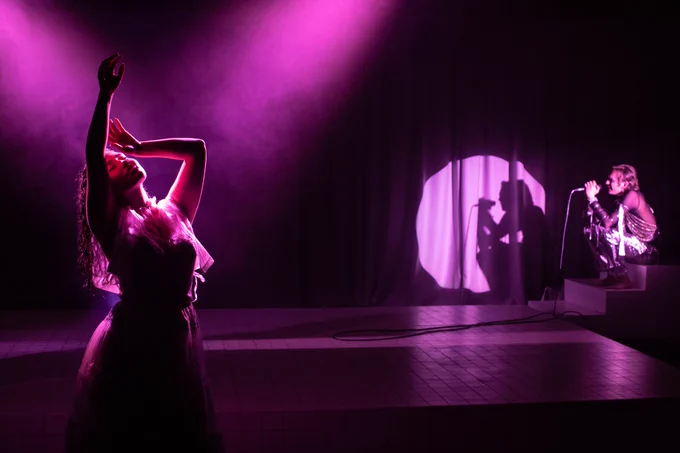Two young performers on a purple lit stage, one with arms raised, another in a circular spotlight singing into a mic
