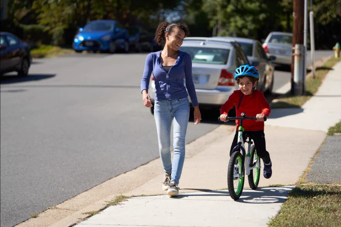 Photograph of a mother and child on their way to school. The child is riding a bicycle and wearing a helmet. The mother is walking beside him.