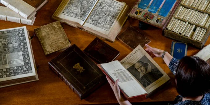 Looking down at a desk full of books from the Special Collections. A person is holding an open book with a picture of William Shakespeare on one page.