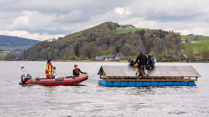 Im Vordergrund: Ein simuliertes schwimmendes Dach auf einem See mit Personen darauf, dem sich von links ein rotes Schlauchboot der Rettungskräfte mit zwei Personen an Bord nähert. Im Hintergrund: Das Seeufer, mit Wald und Wiesen bewachsen, das zu einem Hügel aufsteigt. Der Himmel ist bewölkt. 

Bild: Land Salzburg / Neumayr / Hofer