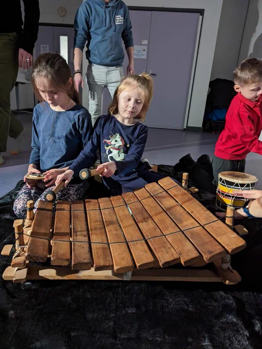 Three children are in a Playground session sitting on a black textured blanket. They are playing with a xylophone and a kalimba and there is a hand holding a djembe (a rope-tuned covered goblet drum)
