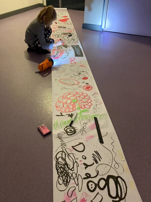 A child is drawing on a long piece of paper whilst sat on the floor. There are a range of drawings in different colour pens and a torch shining on where they are drawing. 