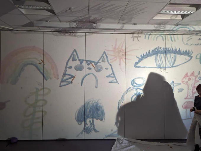 Some drawings from a Playground session are projected onto a white wall. They include a tree, car, rainbow an eye, dinosaur, the sun and more.