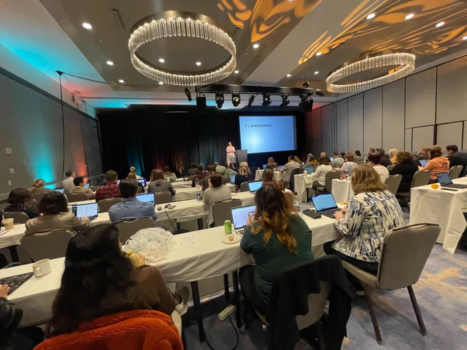 People sitting at tables in a large meeting room, while listening to a presenter and viewing a presentation on a screen.