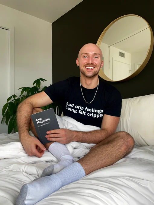 Logan, a white person with a shaved head and short mustache and beard, sits in bed while smiling at the camera. They are wearing white socks and a black shirt that says, “bad crip feelings being felt cripply.” They are holding the book Crip Negativity, which has a solid black cover with white lettering. The bed linens are white.