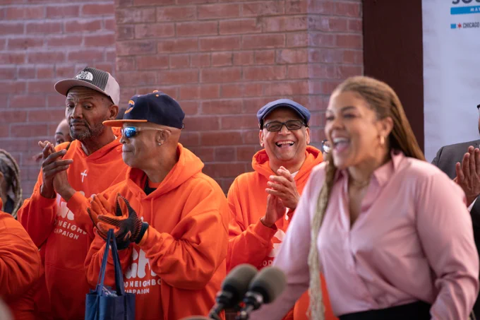 Photo ID: A person is speaking behind a podium and a group of people are cheering behind them. 