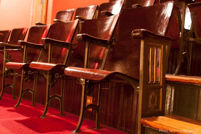 rows of dark-stained wooden theater seats set into red carpet feature wire hat racks beneath